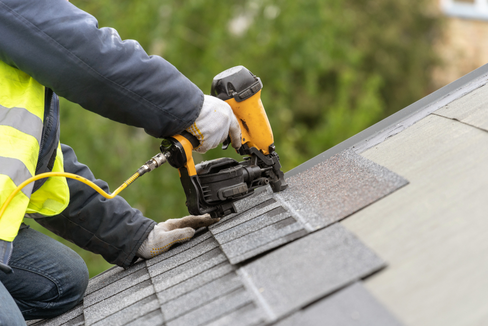 Unrecognizable roofer in uniform using a pneumatic nail gun to lay asphalt or bitumen shingles on a house under construction
